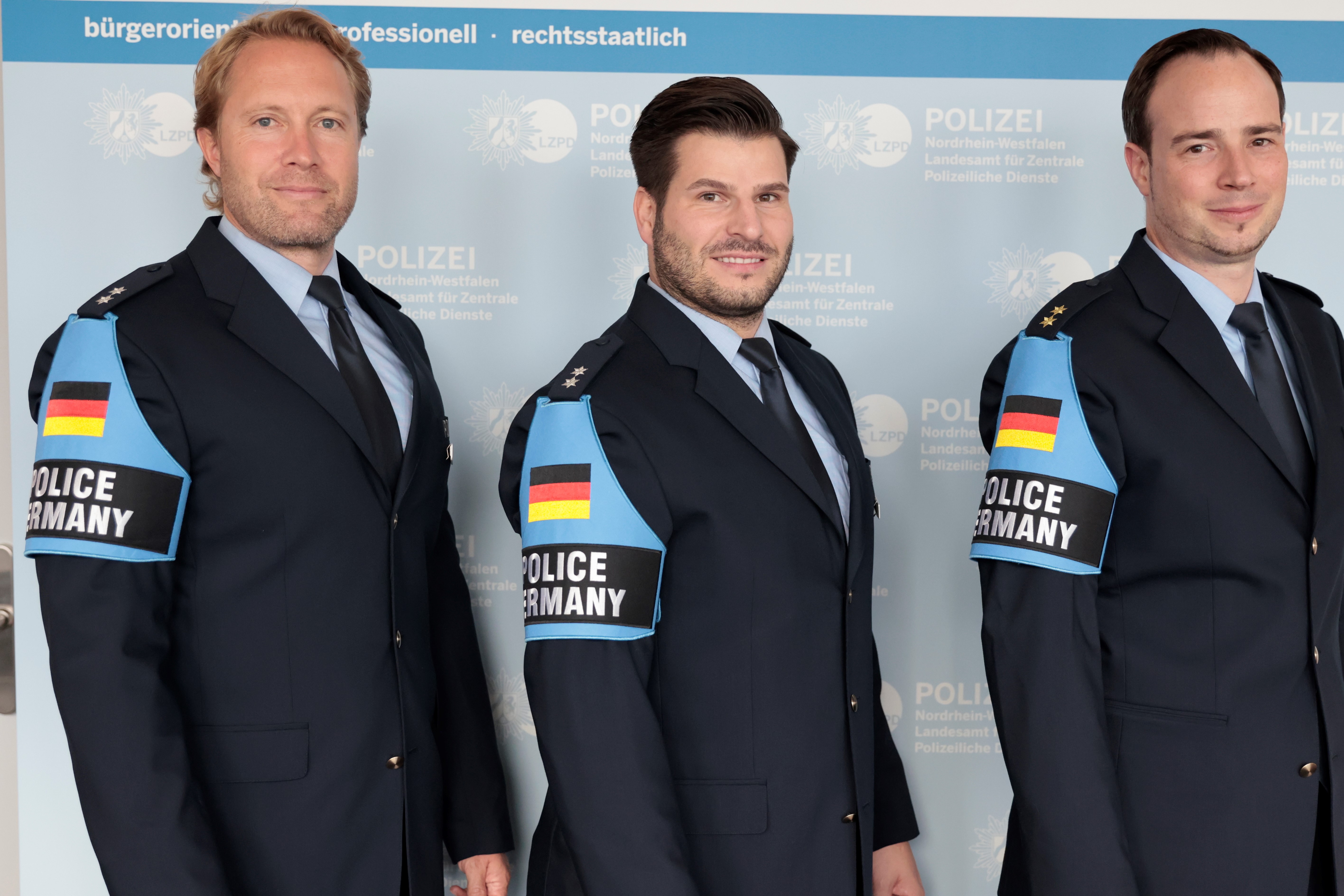 Polizeibeamte stehen nebeneinander. Am Arm tragen sie eine Kennzeichnung mit Deutschlandflagge und der Aufschrift Police Germany.