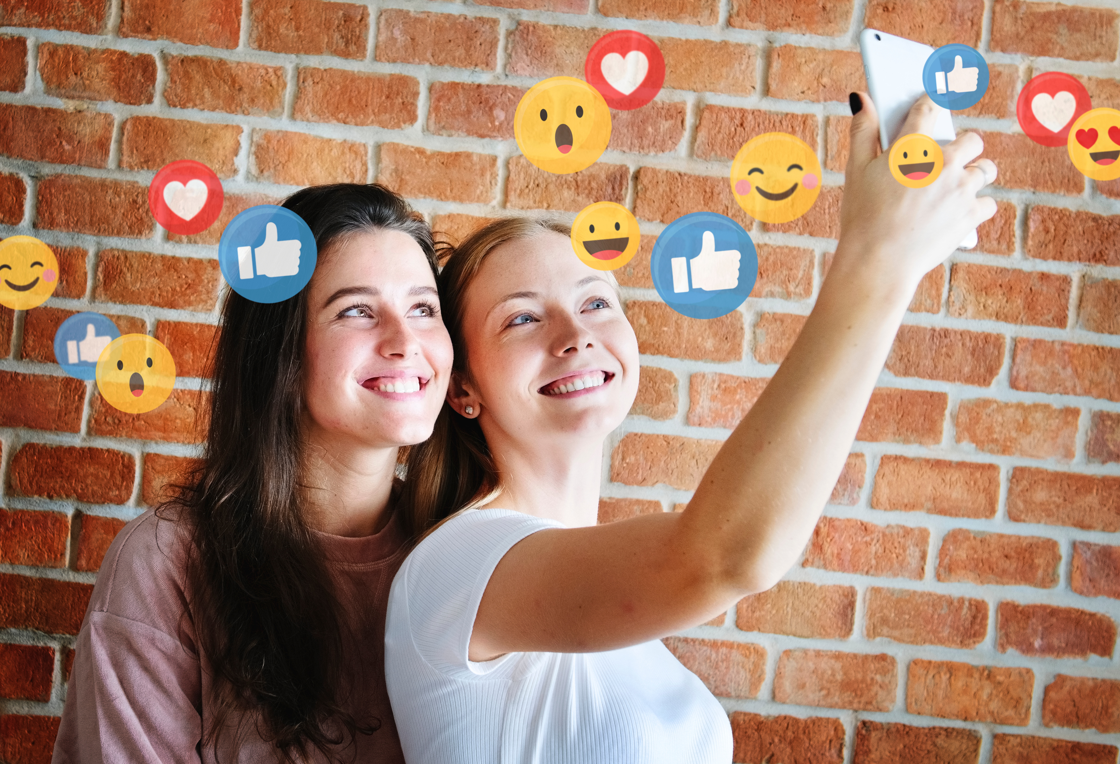 Zwei junge Frauen machen zusammen ein Selfie. Über ihnen sind Social Media Icons wie Emojis Herzen und Daumen hoch zu sehen. Im Hintergrund ist eine Backsteinmauer zu sehen.