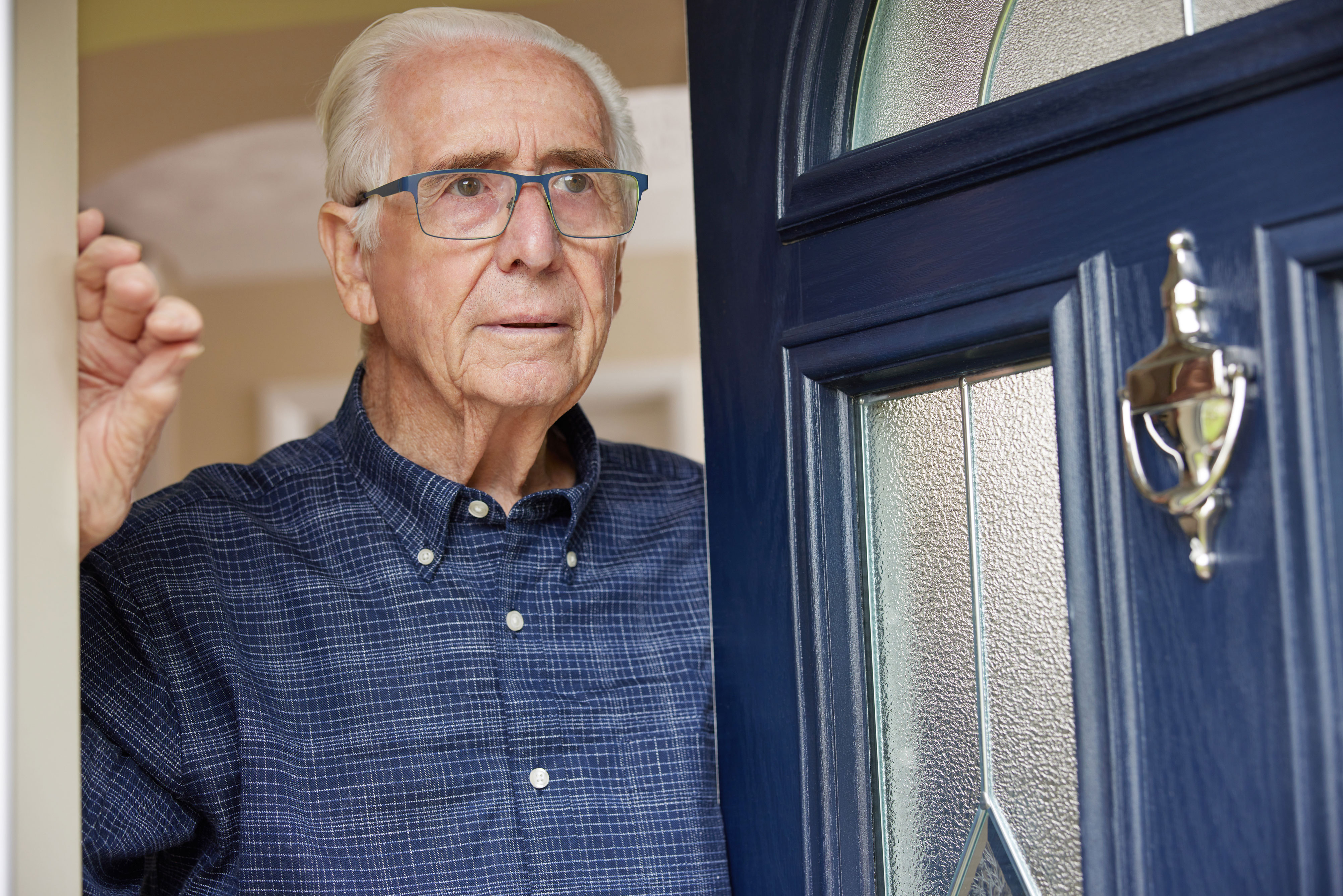 Alter Mann mit blaukariertem Hemd und blauer Brille öffnet die blaue Haustür und blickt nach draußen. Die Haustür hat Fenstereinsätze und einen Türklopfer.
