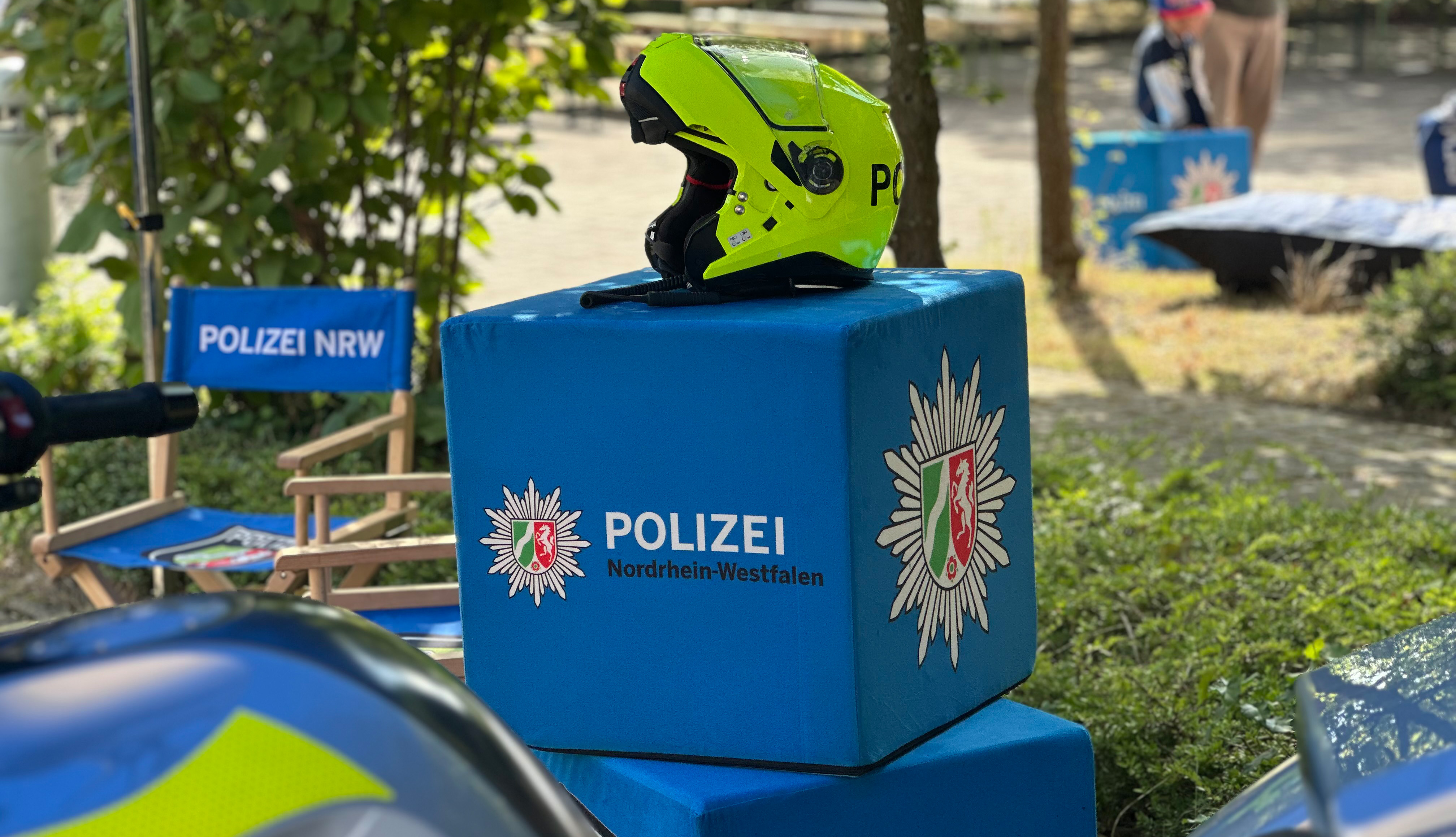 links steht ein Regiestuhl mit der Aufschrift Polizei NRW vor einem Gebüsch. Im Vordergrund ist ein blauer Würfel zu sehen, auf dem ein Polizeistern und Nordrhein Westfalen abgebildet ist. Auf dem Würfel ist ein neongelber Motorradhelm zu sehen. Links unten ist noch ein kleines Stück des Tanks eines Polizeimotorrad in den Farben blau und neongelb zu erkennen.