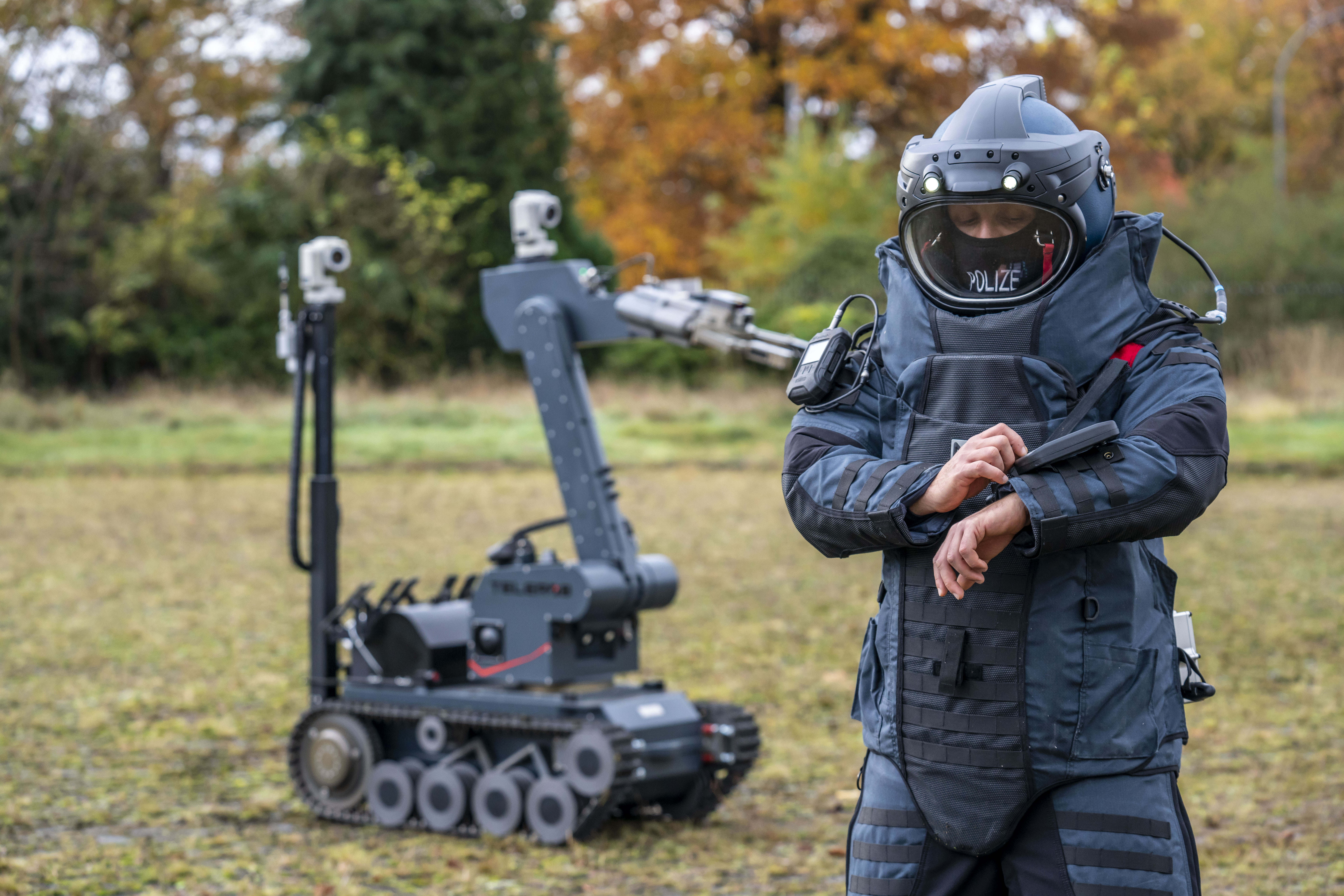 Auf einer Wiese steht ein Sprengstoff-Entschärfer in Schutzkleidung im Vordergrund, der auf eine digitale Anzeige an seinem linken Arm schaut; im Hintergrund steht ein Sprengstoffroboter