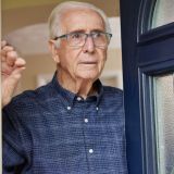 Alter Mann mit blaukariertem Hemd und blauer Brille öffnet die blaue Haustür und blickt nach draußen. Die Haustür hat Fenstereinsätze und einen Türklopfer.