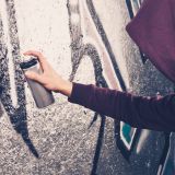 Eine Person mit auberginefarbenem Hoodie sprüht ein Graffiti an eine Wand