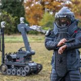 Auf einer Wiese steht ein Sprengstoff-Entschärfer in Schutzkleidung im Vordergrund, der auf eine digitale Anzeige an seinem linken Arm schaut; im Hintergrund steht ein Sprengstoffroboter