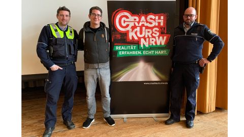 MaximeNoise  und zwei Polizisten stehen vor einem Plakat. Auf dem Plakat steht Crash Kurs NRW. 