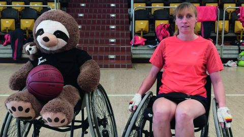 In the foreground, policewoman Sandra Winkler sits in a sports wheelchair. Next to her is another wheelchair in which a cuddly toy mascot is sitting. They are in a sports hall. Empty rows of seats can be seen in the background.
