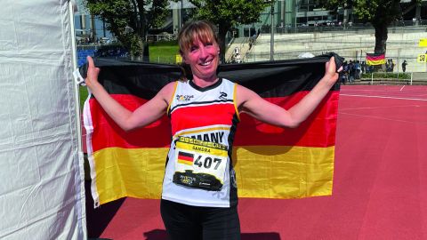 Policewoman Sandra Winkler stands on a sports field wearing a Germany jersey. She is holding a German flag over her back. Trees and a section of the Düsseldorf Arena can be seen in the background