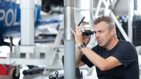 David Horn sits on the floor of the hangar and measures the depth of scratches discovered on a rotor mast. He is wearing dark work clothes. A police helicopter and a lifting platform can be seen in the background.
