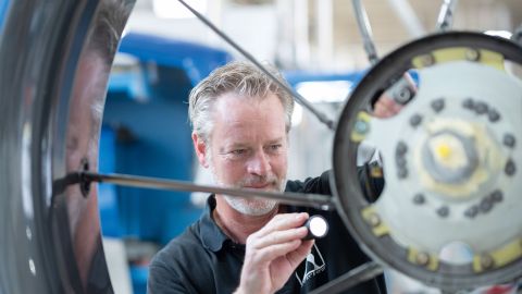 Mechanic Marc Dietel inspects the tail rotor of an Airbus H145 using a flashlight. Large pieces of equipment can be seen blurred in the background.