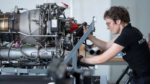 Inspector Stefanie Bohr examines a large engine consisting of many parts. A white wall can be seen in the background.