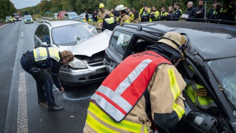 Ein Mitglied der Feuerwehr untersucht ein Unfallauto. Eine Polizistin markiert mir Kreide die Endposition eines anderes Unfallautos. Im Hintergrund sind weitere Beschäftige der Polizei und Feuerwehr zu sehen, sowie Fahrzeuge.