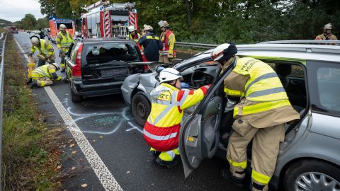 Feuerwehrleute stabilisieren zwei Unfallsautos mit einem Stabblocksystem. Im Hintergrund sind weitere Feuerwehrleute und Fahrzeuge zu sehen.