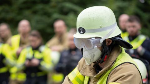 Ein Mitglied der Feuerwehr schlägt mit einem Notfallhammer die Scheibe eines Unfallautos ein. Im Hintergrund sind Weitere Feuerwehrleute und Polizeikräfte zu sehen.