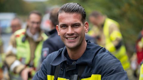 Christoph Pütz, Einsatztrainer der Feuerwehr Aachen ist im Fordergrund forntal zu sehen. Er lächelt in die Kamera. Er befindet sich bei einer Einsatzübung auf einer Autobahn. Im Hintergrund sind weitere Feuerwehrkräfte zu sehen.