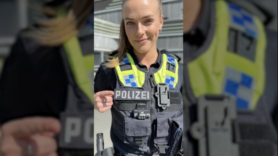 Eine uniformierte Polizistin zeigt lächelnd mit dem Zeigefinger auf die Aufschrift "Polizei" auf ihrer Weste.