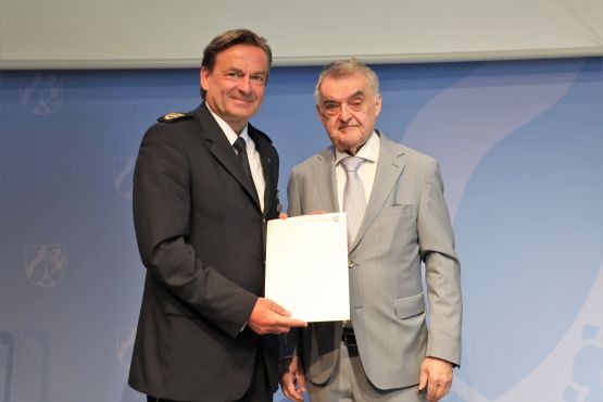 Thomas Roosen und Innenminister Herbert Reul stehen nebeneinander. Roosen hält seine Ruhestandsurkunde in der Hand.