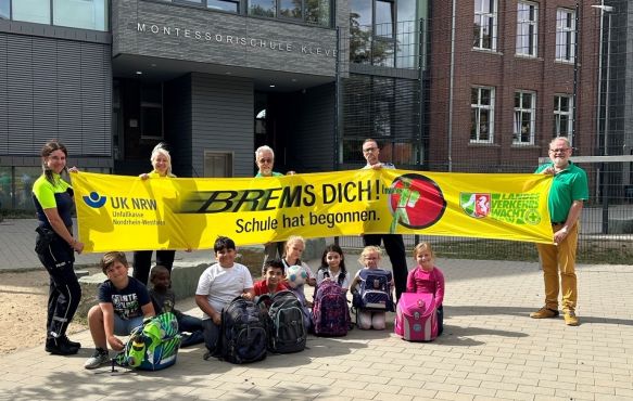 auf dem Schulhof der Montessori Schule Kleve stehen mehrere Polizeibeamter und Mitarbeiter der Verkehrswacht und halten ein Banner der Aktion "Brems Dich" hoch. Vor dem Banner sitzen mehrere Schülerinnen und Schüler der Schule