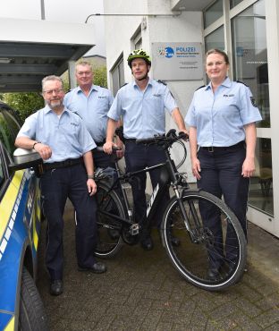 Drei Polizeibeamte und eine Polizeibeamtin mit Fahrrad und Streifenwagen vor dem Eingang einer Polizeiwache